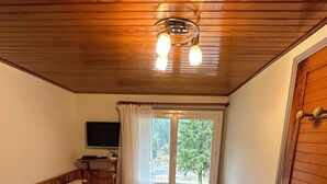 Quarto com teto de madeira, cama com roupa de cama listrada, janela com cortinas, televisão e radiador.