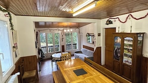 Uma sala de jantar com painéis de madeira, uma mesa comprida com bancos, um armário de madeira com quadros e vista para o exterior através de grandes janelas.