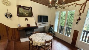 Um quarto aconchegante com teto de madeira, lareira, mesa de jantar com cadeiras, televisão e uma grande janela com vista para árvores.