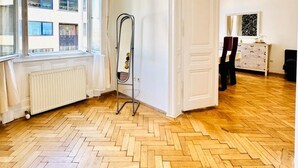 A spacious room with a wooden parquet floor, a radiator, a metal drying rack, and a view of a building through large windows. - Cozy 1BR and Living Room Apartment (Vienna)