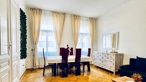 A spacious living area with a wooden herringbone floor, a dining table with chairs, a sofa, a cabinet, and a large window with curtains. - Cozy 1BR and Living Room Apartment (Vienna)