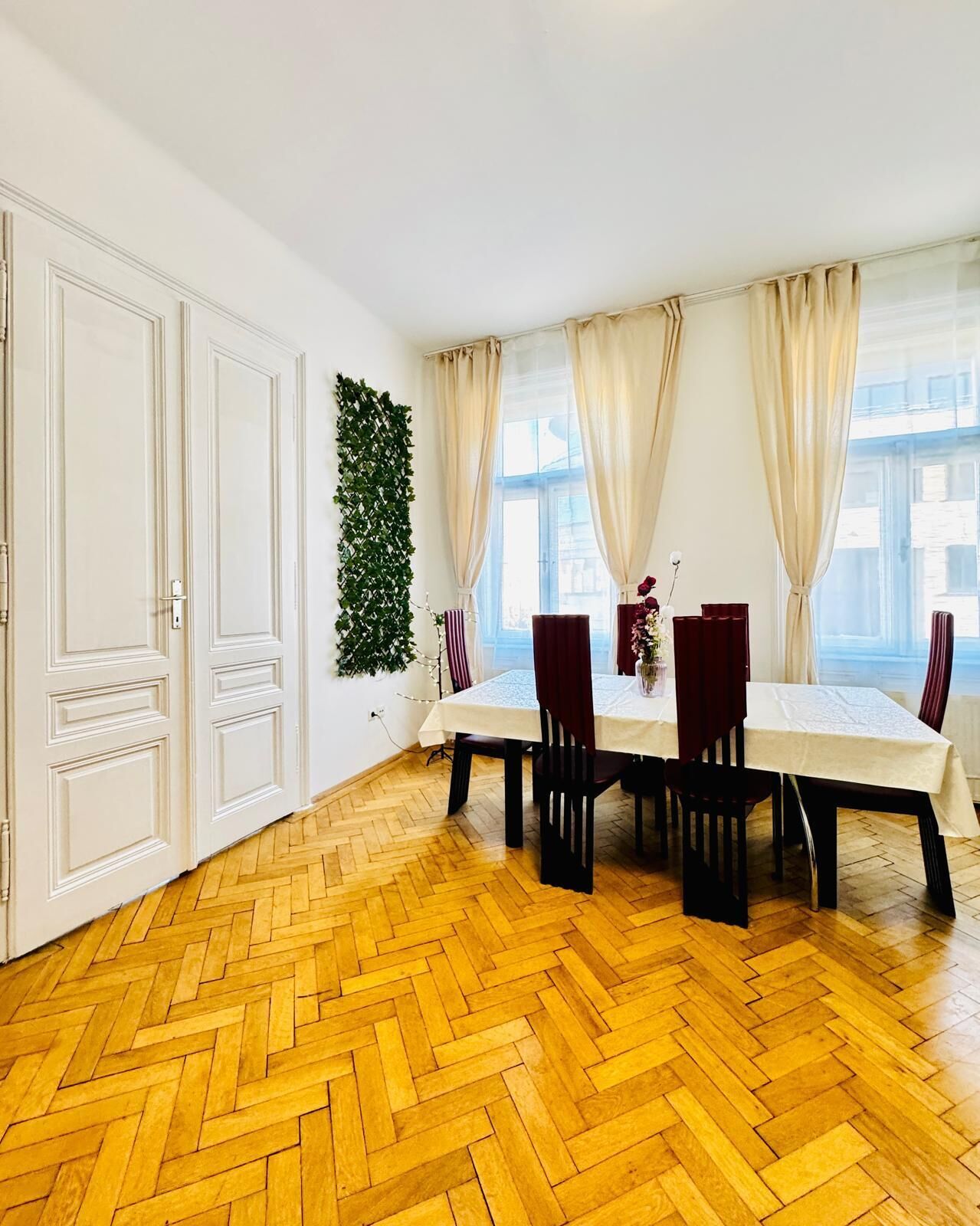 Un espace repas doté d’un parquet en bois à motifs chevrons, de murs blancs et d’une grande fenêtre agrémentée de rideaux beiges.