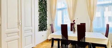 Un espace repas doté d’un parquet en bois à motifs chevrons, de murs blancs et d’une grande fenêtre agrémentée de rideaux beiges.