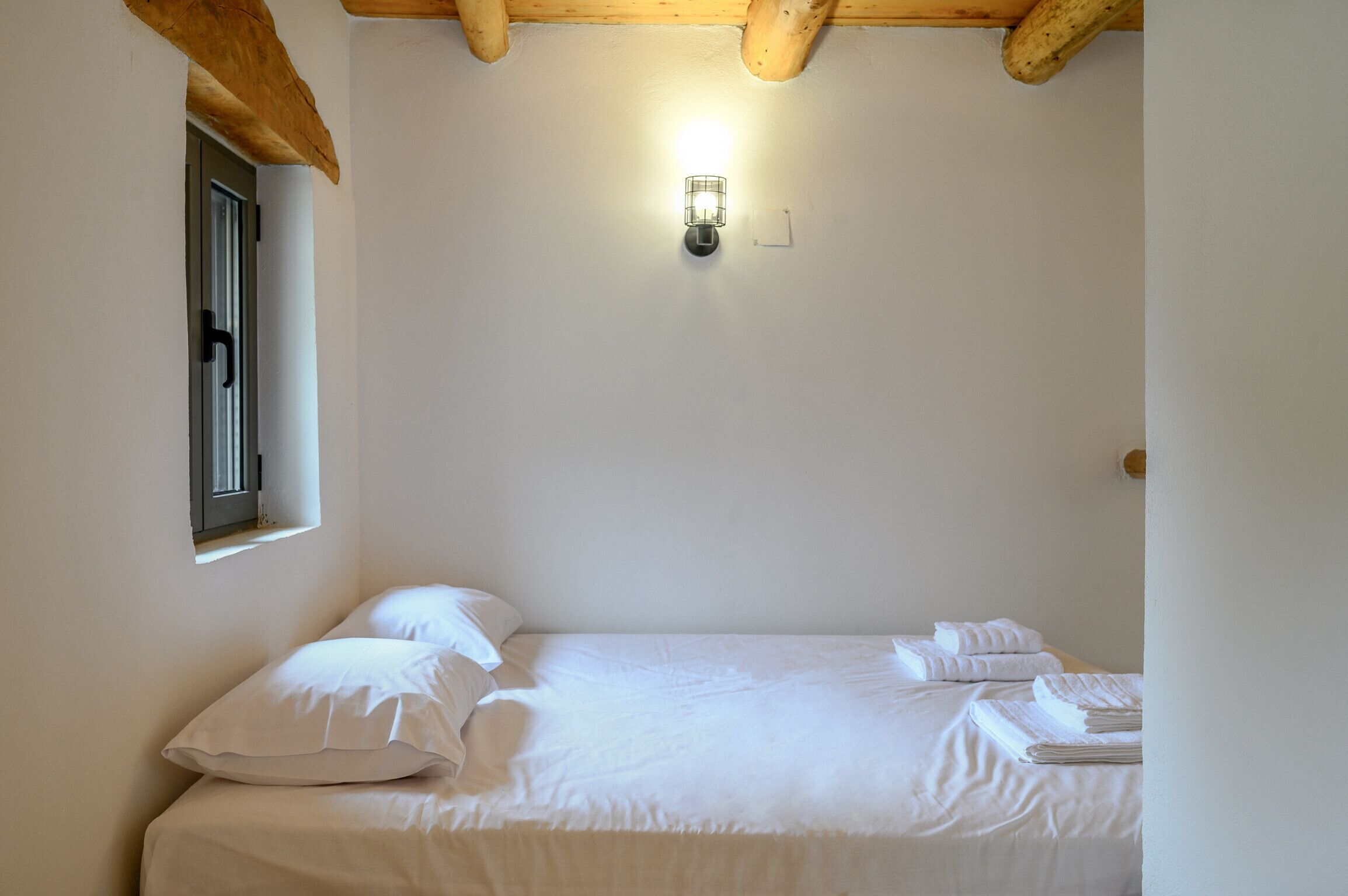 A single bed with white linens and pillows, a small window with a view, and a wall-mounted light fixture.
