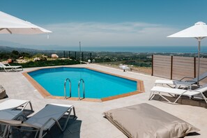 Ein rechteckiger Pool mit Metallleiter, umgeben von Liegestühlen und Sonnenschirmen, mit Blick auf eine malerische Meerlandschaft.