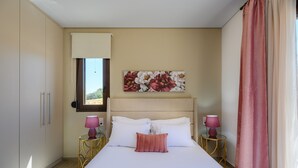 A bedroom with a bed, two bedside tables with lamps, a window with curtains, and a floral artwork on the wall. - Jason Panorama Villa 1 (Rethymno)