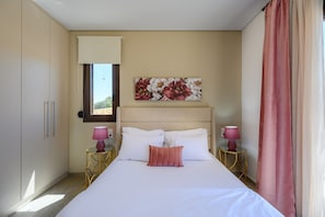 A bedroom with a bed, two bedside tables with lamps, a window with curtains, and a floral artwork on the wall. - Jason Panorama Villa 1 (Rethymno)