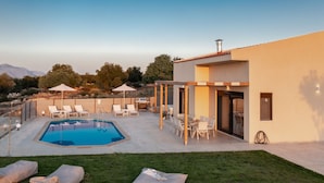 Un área de piscina moderna al aire libre con sillones de descanso y sombrillas, rodeada de un césped cuidado y un edificio con amplios ventanales.