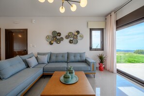 A modern living room with a sectional sofa, a wooden coffee table, decorative wall art, and a large window with a view. - Jason Panorama Villa 2 (Rethymno)