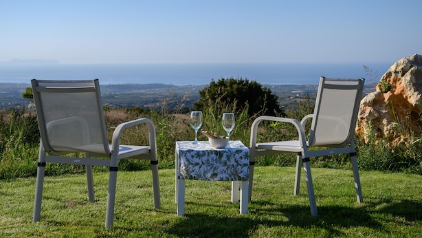 Dos sillas blancas y una mesa con mantel floreado, ubicadas en un césped con vista al mar y un paisaje distante.