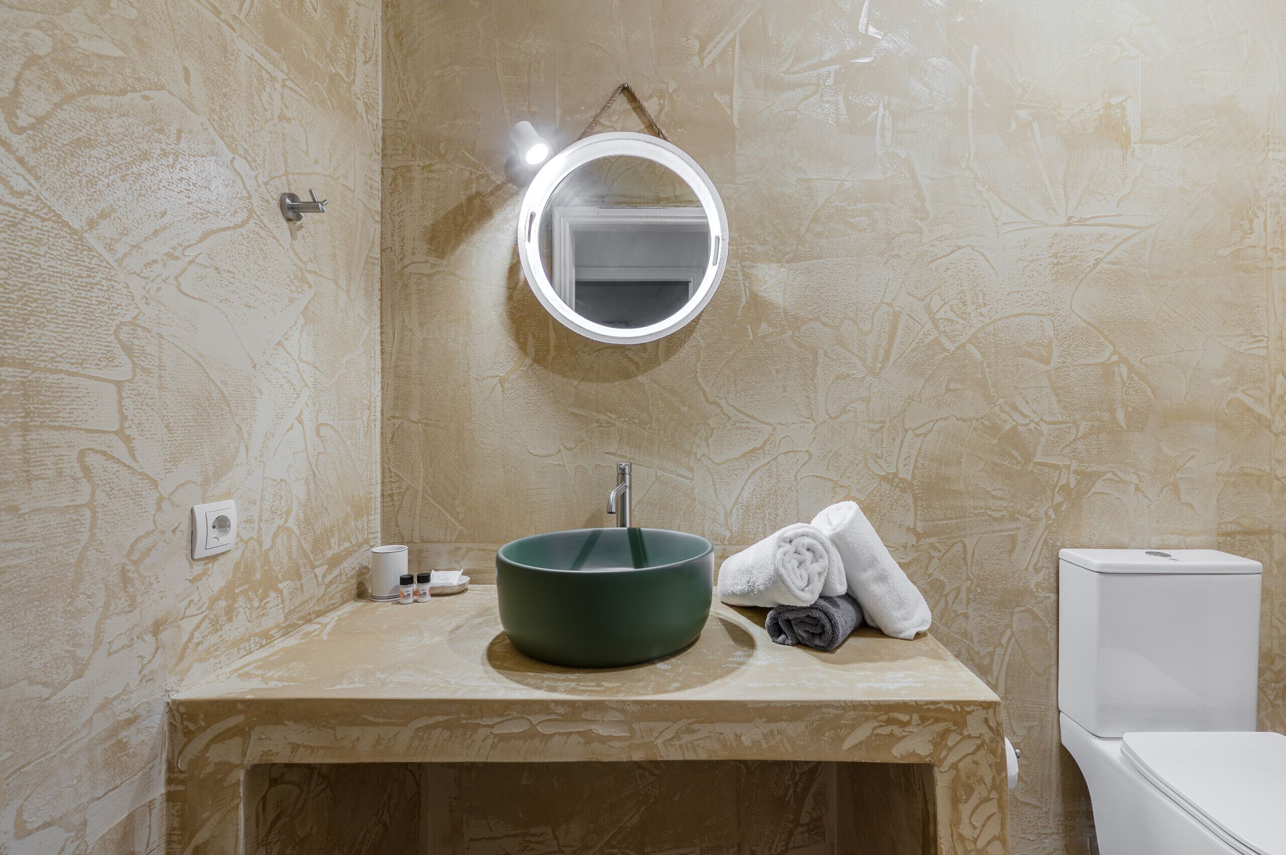Un baño con un espejo redondo, un lavabo verde, un inodoro y toallas sobre el mostrador.