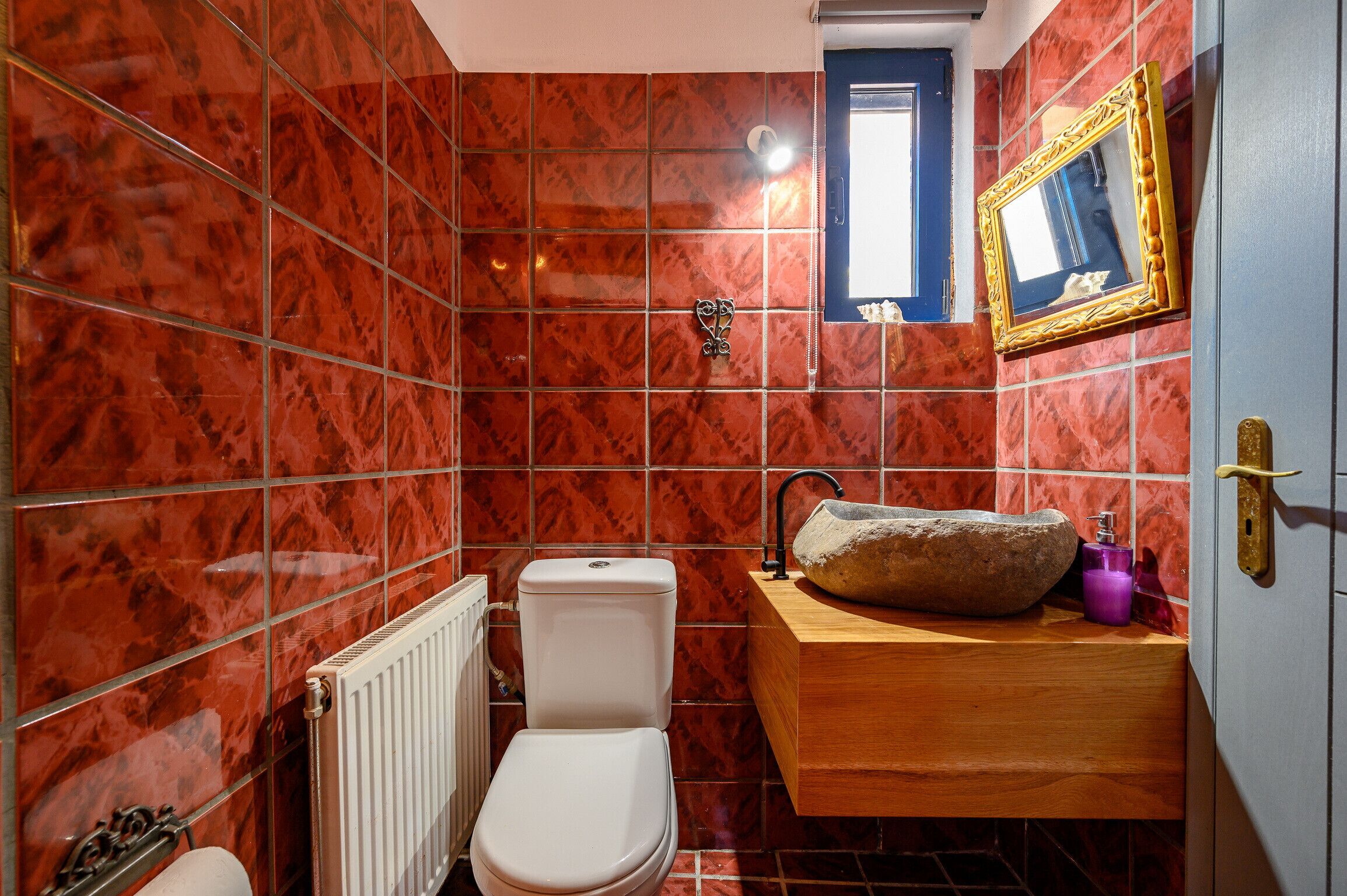 Een badkamer met rode marmeren tegels, een wit toilet, een houten wastafelmeubel met een stenen wastafel, een spiegel met gouden lijst en een raam boven de wastafel.