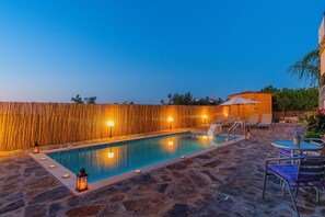 Ein Pool im Freien mit Steinplattenboden, umgeben von Liegestühlen und Sonnenschirmen, mit einem Holzzaun im Hintergrund.