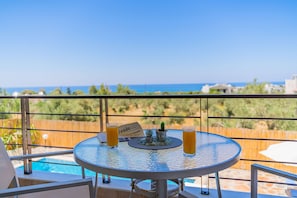 Ein runder Tisch mit zwei Orangensaftgläsern, einem kleinen Topfpflänzchen und einer Speisekarte auf einem Balkon mit Blick auf einen Pool und das Meer.