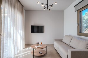 A modern living room with a sofa, a round wooden coffee table with a book and a glass of orange juice, a flat-screen TV on the wall, and a large window with curtains. - Meli & Gio Villa 1 (Rethymno)
