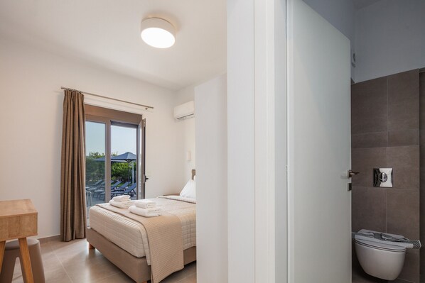 A hotel room with a bed, a wooden table, a window with curtains, and a view of a pool and mountains. - Angeliana Villa (Mylopotamos)