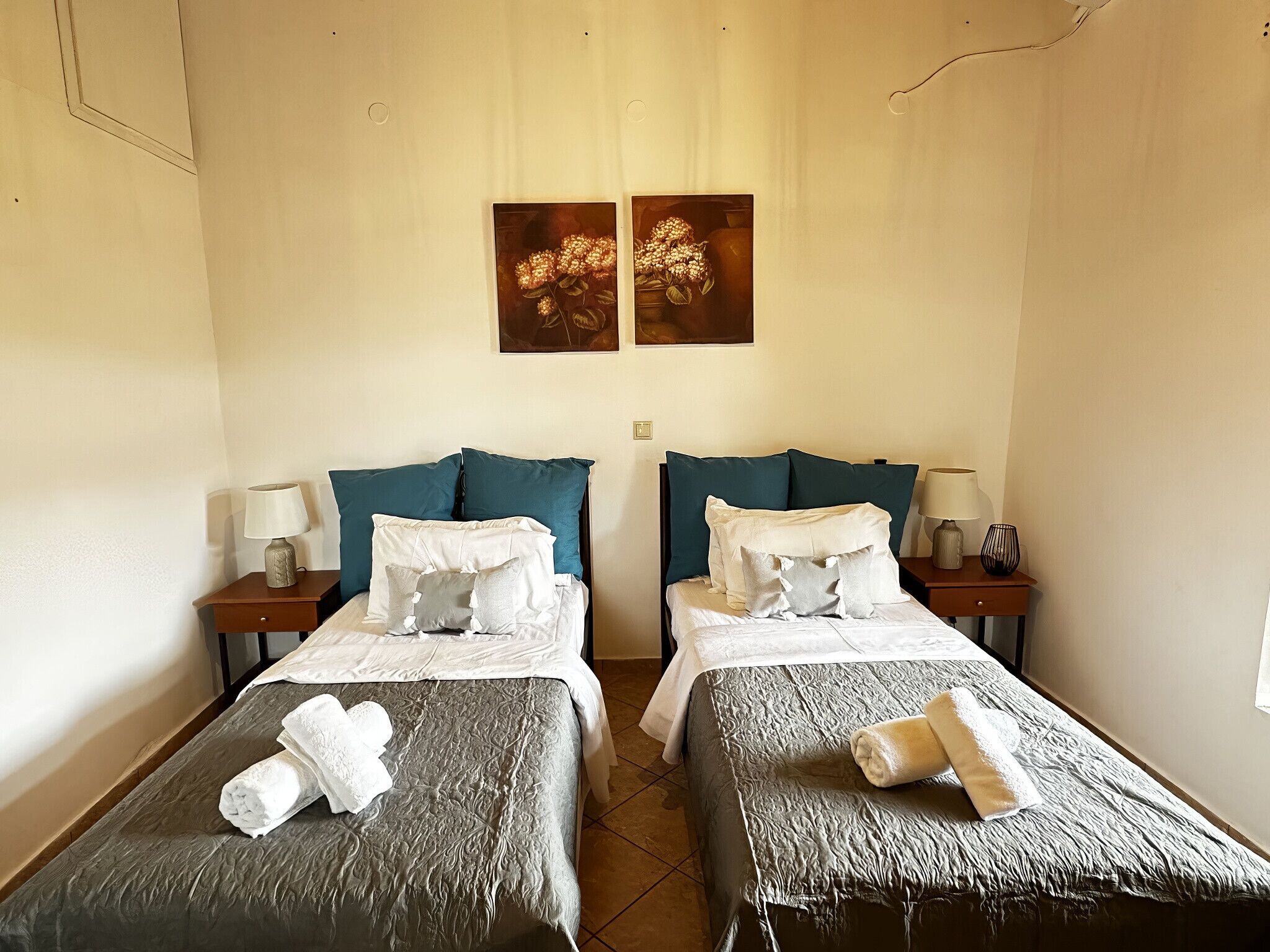 Quarto de hotel com duas camas de solteiro, cada uma com uma mesa de cabeceira, luminária e um quadro pendurado acima.