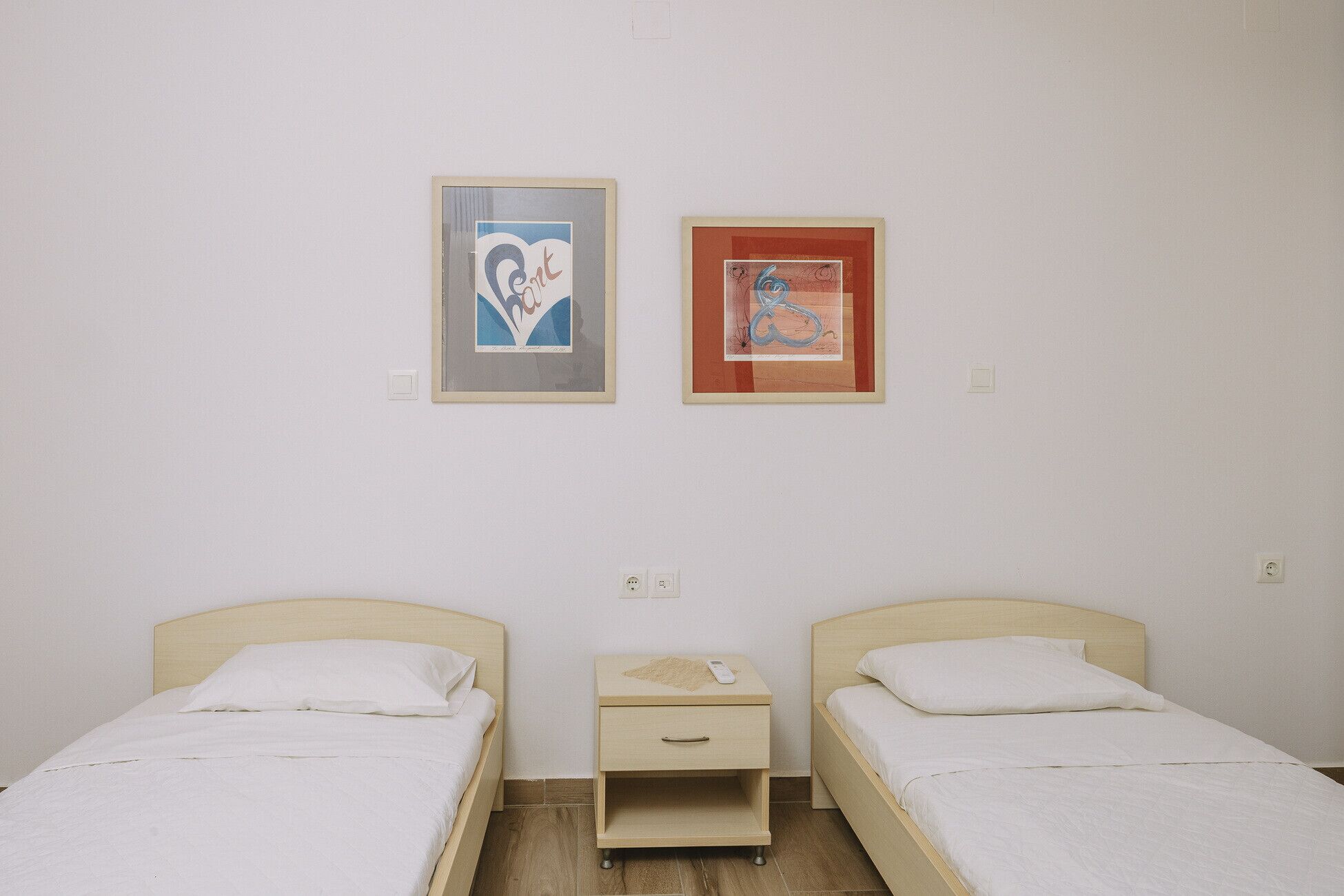 A room with two single beds, a small bedside table with a lamp, and two framed artworks on the wall.