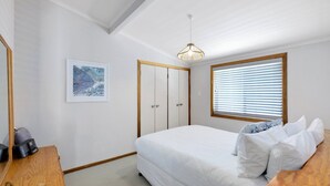 A bedroom with a bed, a wooden dresser, a framed painting, a wardrobe, and a window with blinds. - Upper Oceanside (AVOCA BEACH)
