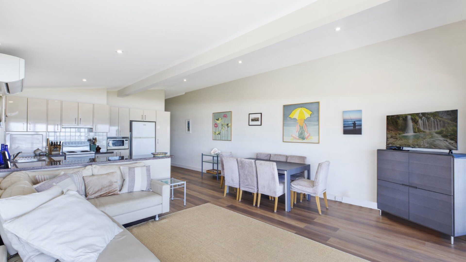 A modern living room with a sofa, dining table, and chairs, a kitchen area with white cabinets and appliances, and artwork on the walls.