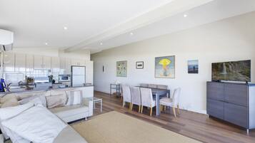 A modern living room with a sofa, dining table, and chairs, a kitchen area with white cabinets and appliances, and artwork on the walls.