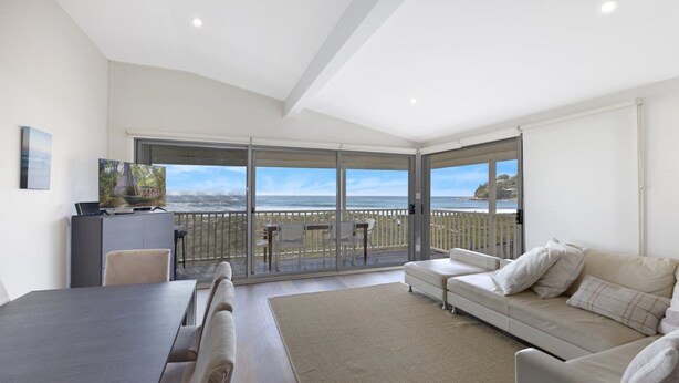 A spacious living area with a balcony overlooking the sea, a dining table, and a sofa. - Upper Oceanside (AVOCA BEACH)
