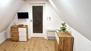 Una habitación pequeña y estrecha con techo inclinado, un armario de madera con cajones, una pequeña mesa con una planta y un televisor montado en la pared.