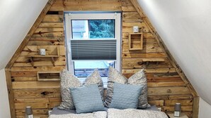 Un dormitorio tipo loft con techo inclinado, vigas de madera, una cama grande con ropa de cama estampada y una ventana con persianas.