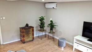 Una sala de estar con piso de madera, un televisor sobre un mueble blanco, dos plantas en macetas, un taburete pequeño y un aparador de madera antiguo.