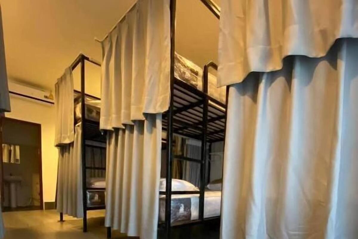 Una fila de literas con cortinas blancas, suelo de madera y una puerta al fondo.