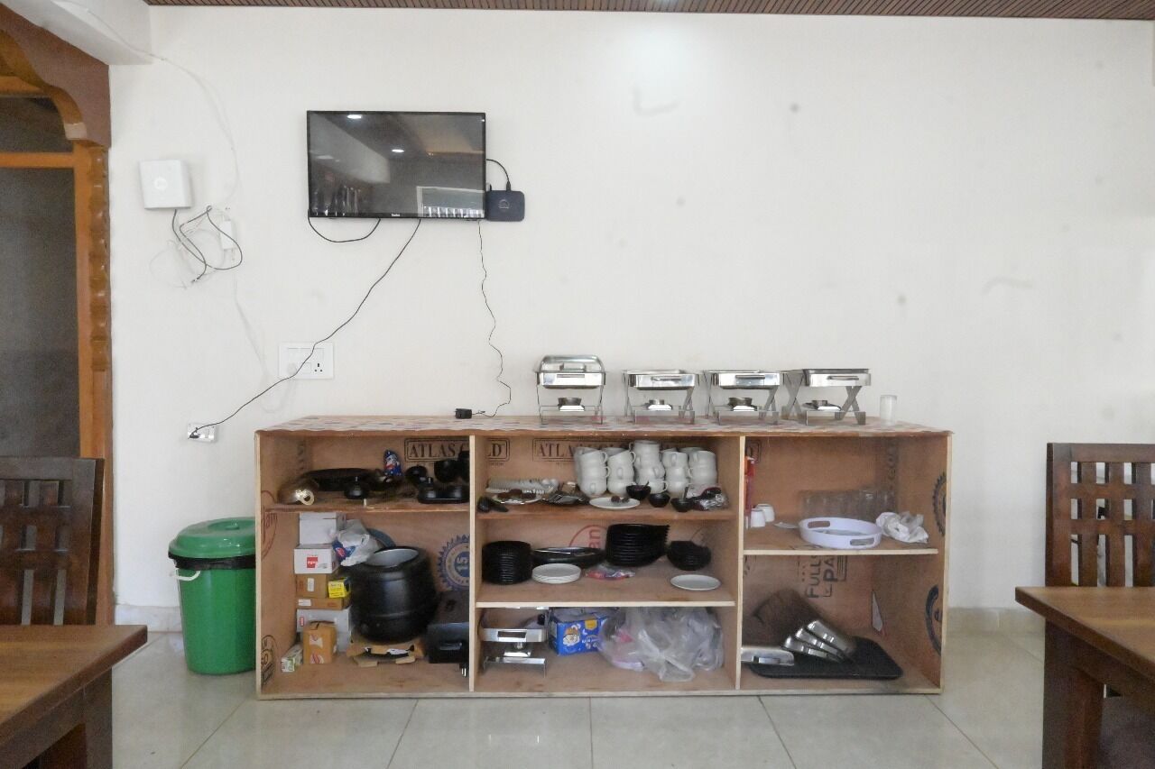 A room with a wooden cabinet containing kitchenware, a television mounted on the wall, and a green trash bin.