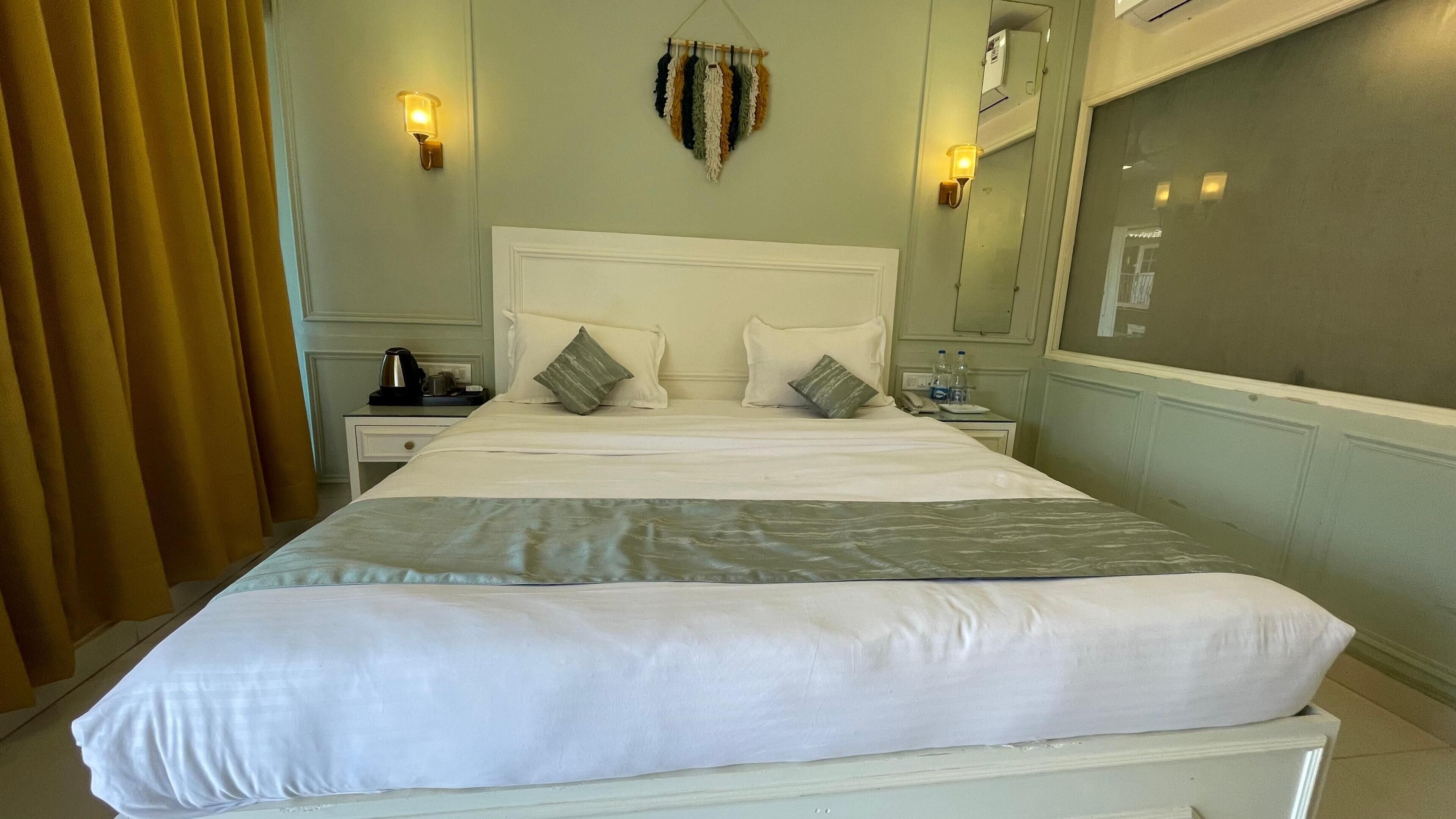 A hotel room with a bed, bedside tables, a nightstand, a mirror, and a wall-mounted light fixture.