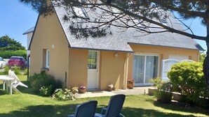 Une maison avec un toit en pente, une porte d’entrée et une porte-fenêtre coulissante. On trouve deux chaises de jardin et une petite table dans le jardin.