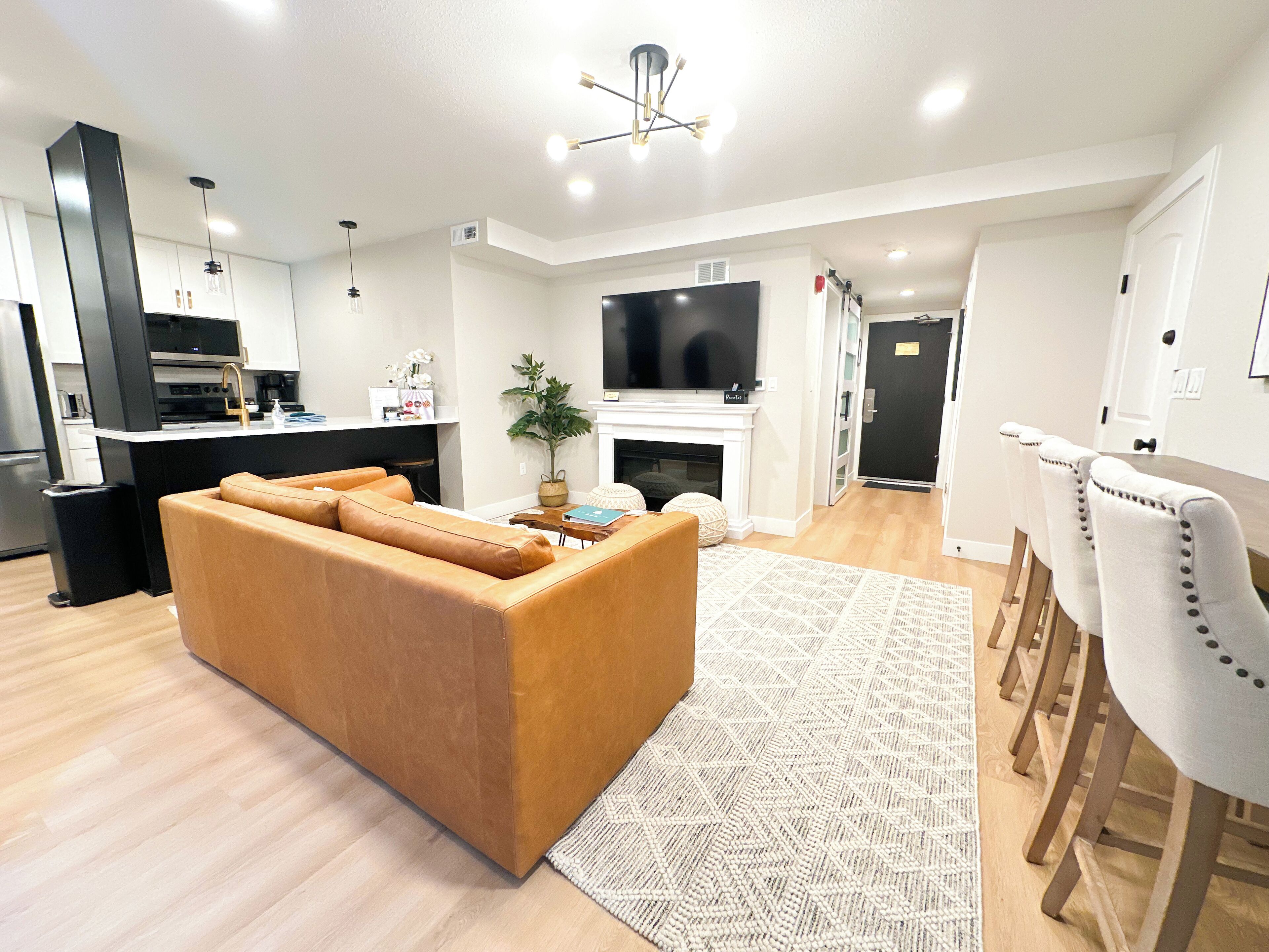 Una sala de estar moderna con un sofá, un comedor con sillas, una cocina con televisión y una chimenea.