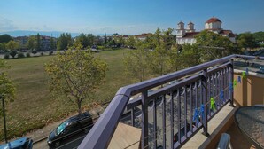 A balcony with a view of a church, a grassy field, and parked cars. - Welcome to Double Room – Olympic Studios by SDR (Paralia)
