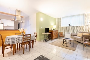 A modern living room with a sofa, a dining table with chairs, a TV, and a kitchen area with cabinets and a stove. - Renovated Apartment in the Heart of Pont-l’Abbé – River View, Close to the Ocean (Pont l'abbé)