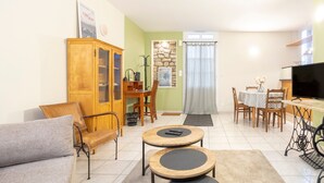 Een woonkamer met een bank, twee ronde tafels, een televisie, een eettafel met stoelen, een houten kast en een stenen muur.