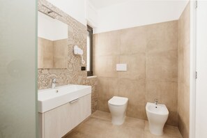 A modern bathroom with a white sink, toilet, and bidet, beige tiles, and a patterned backsplash. - B&B Castello Otranto (Otranto)