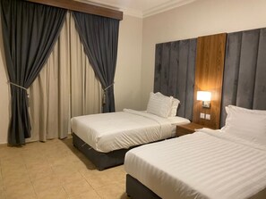 A hotel room with two beds, a wooden headboard, a nightstand with a lamp, and a window with curtains. - Manazel Al Khaleej Hotel Apartments (Dammam)