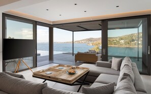 Ein modernes Wohnzimmer mit einem großen Glasfenster, das auf einen Balkon mit Blick auf ein Gewässer und eine hügelige Landschaft führt.