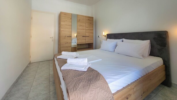 A bedroom with a wooden bed, a wardrobe, and a nightstand with a lamp. - ARETI Deluxe Two - Bedroom Suite n. 101 By SDR (Olimpiaki Akti)
