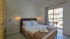 A hotel room with a large bed, two bedside lamps, a window with a view of a building, and a wooden door. - ARETI Deluxe Two - Bedroom Suite n. 101 By SDR (Olimpiaki Akti)