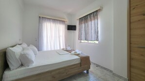 A bedroom with a wooden bed, a window with curtains, a TV mounted on the wall, and a wooden wardrobe. - ARETI Deluxe Two - Bedroom Suite n. 101 By SDR (Olimpiaki Akti)