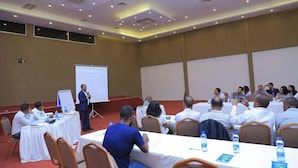 A conference room with a speaker at the front, attendees seated at tables, and a presentation screen.
