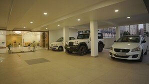 Un moderno concesionario de autos con tres vehículos expuestos, una zona de recepción con mostrador y plantas, y vistas de edificios al exterior.