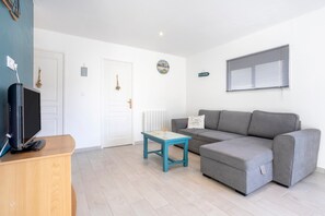 A living room with a grey sofa, a wooden TV stand, a small table, and a window with blinds.