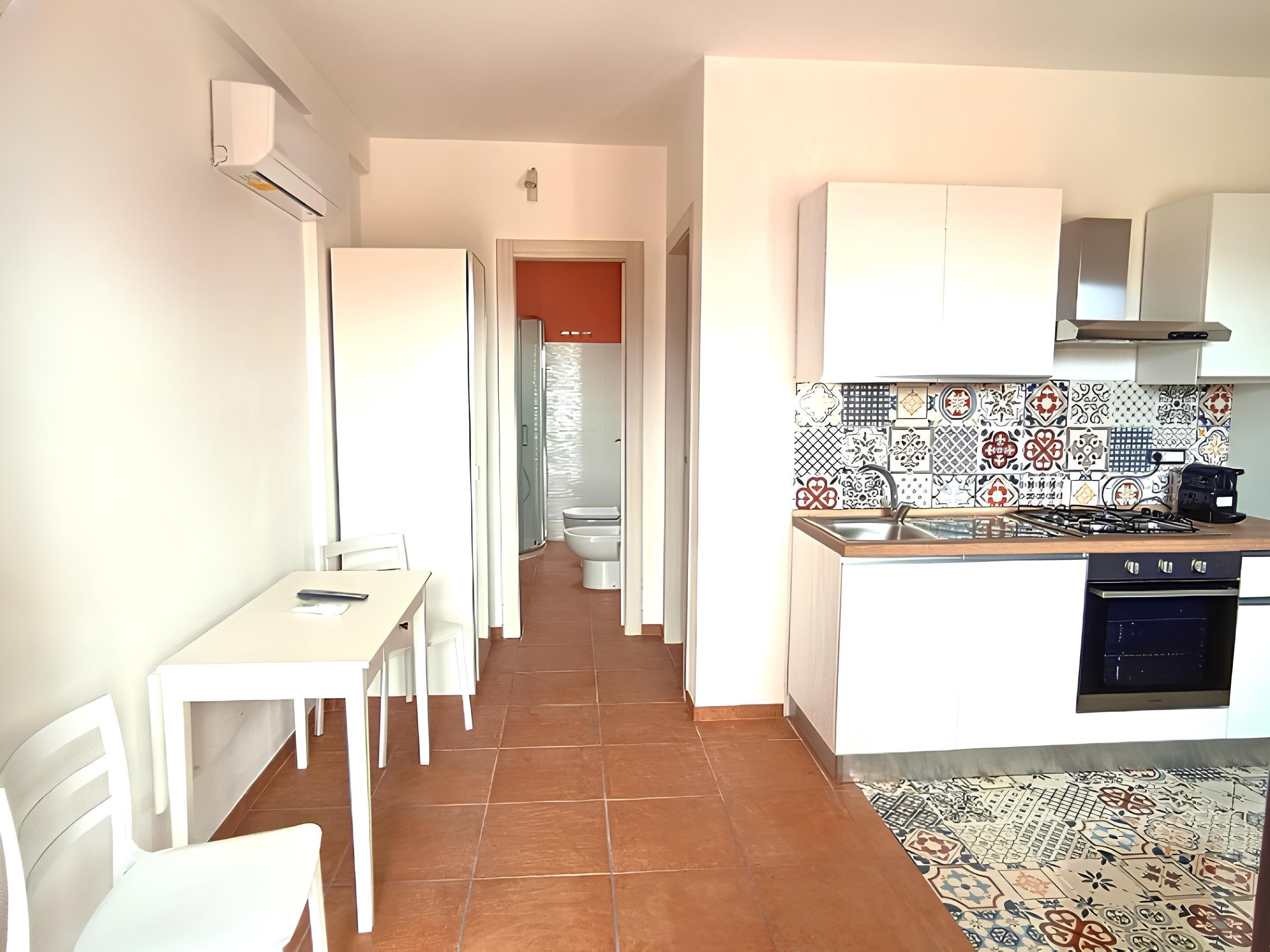 Una cucina compatta con mobili bianchi, zoccolo in piastrelle e zona pranzo con tavolo e sedie.