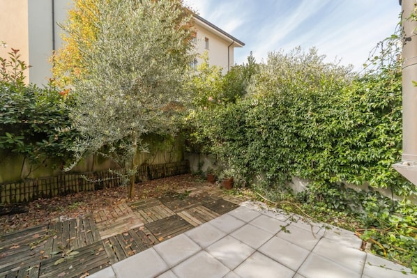 A courtyard with a wooden deck, a tiled patio, and dense greenery including a tree and bushes.