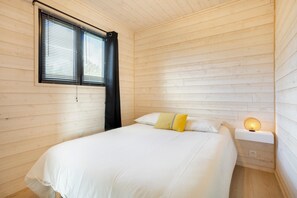 Ein Zimmer mit Holzwänden, einem Bett, einem Nachttisch mit Lampe und einem Fenster mit Jalousien.