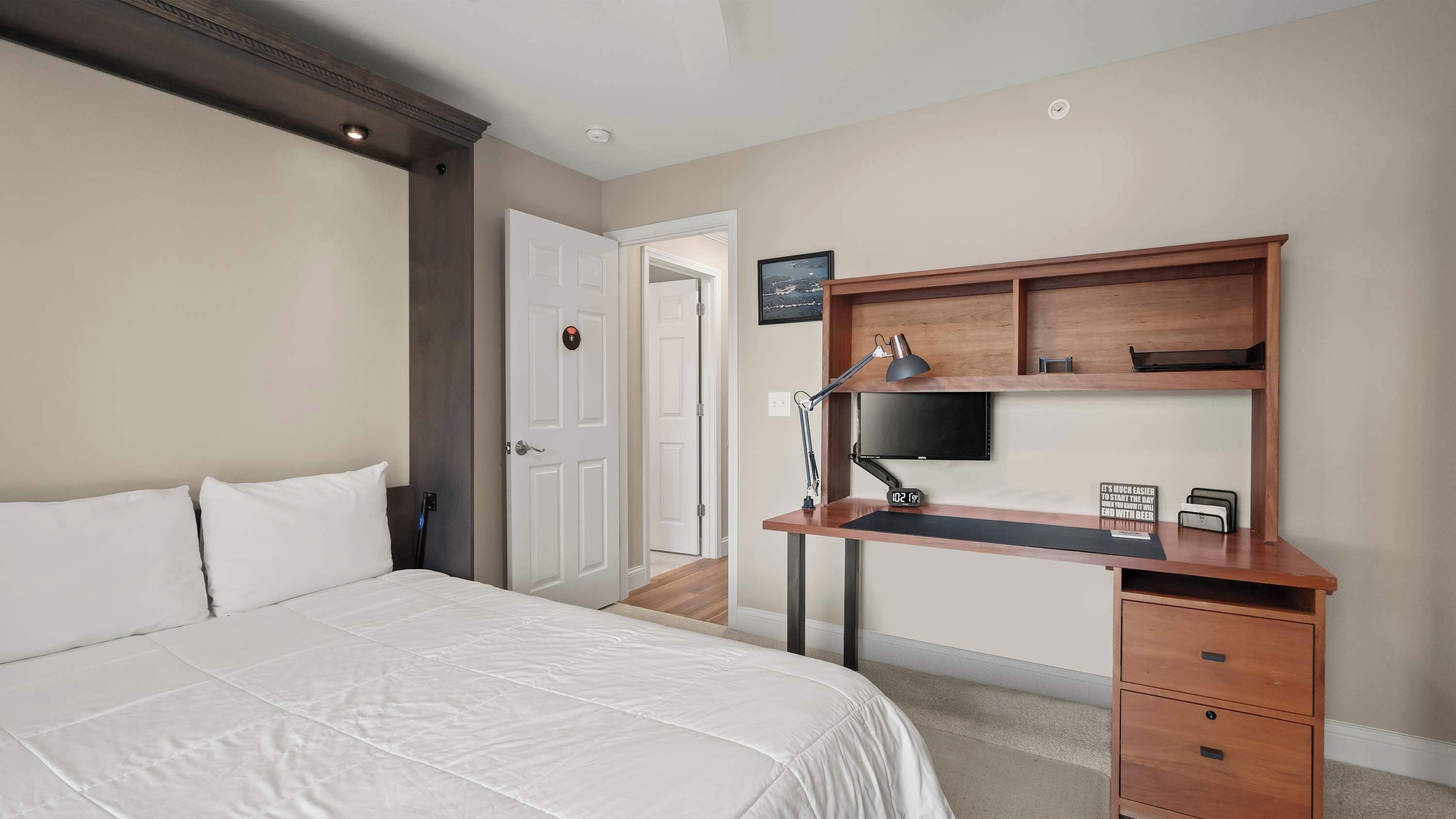 A hotel room with a bed, a desk with a computer monitor, a lamp, and a clock.